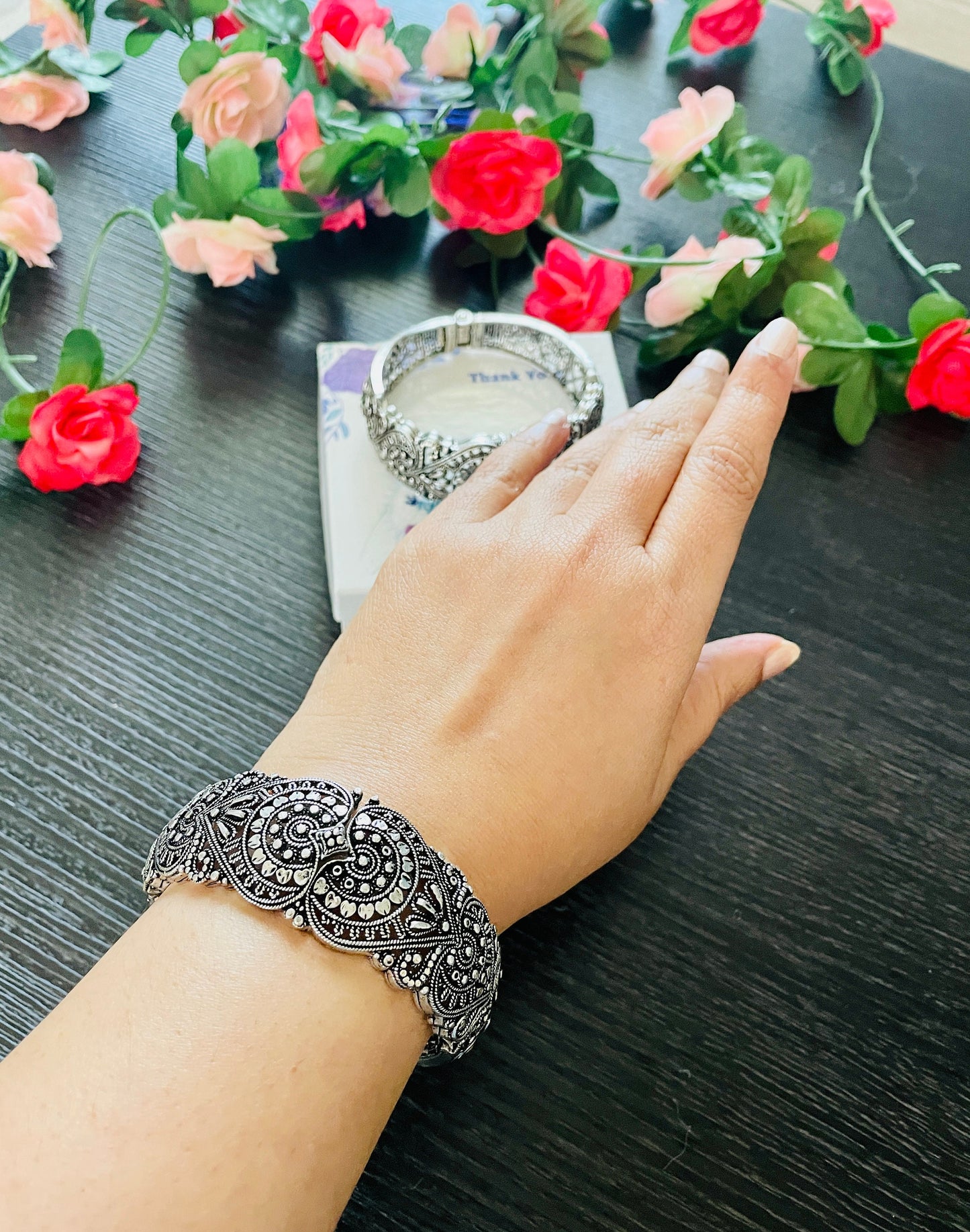A silver-toned cuff bracelet with an ornate, detailed design featuring floral and geometric patterns. The bracelet is on a wrist, showcasing its intricate craftsmanship. A second bracelet is visible in the background.