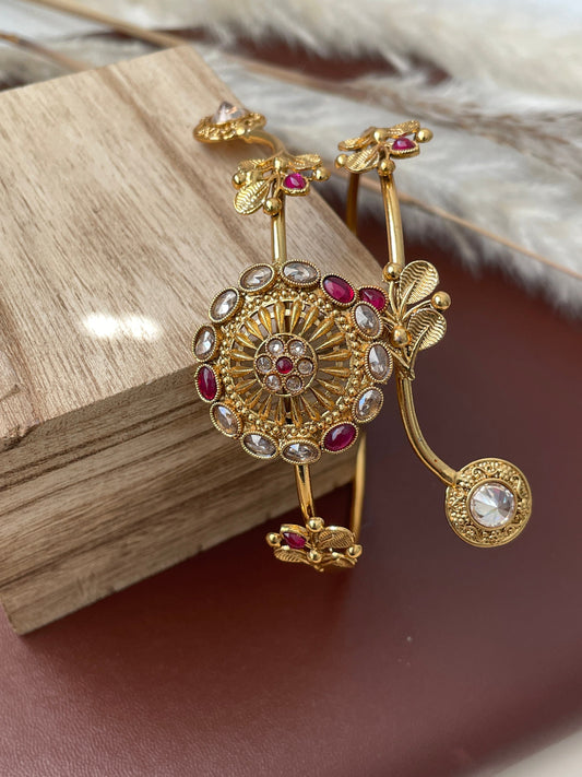 Gold plated armlet with red and white stones. The armlet is decorated with intricate floral designs and has a large, round centerpiece with a circular pattern of white stones and a red stone in the center.