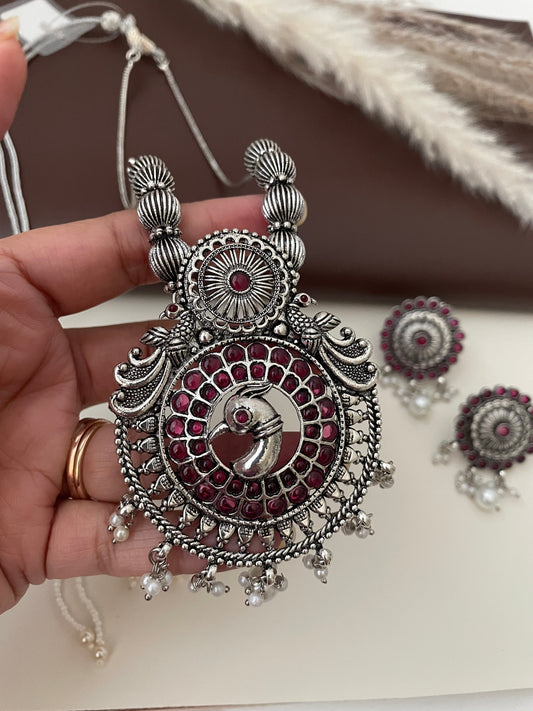 A silver necklace with a pendant featuring a peacock design with ruby kemp stones. The pendant is surrounded by a circle of smaller pink stones. The necklace has a silver chain and a silver tassel with small white pearls. The necklace is paired with matching earrings with a similar design.