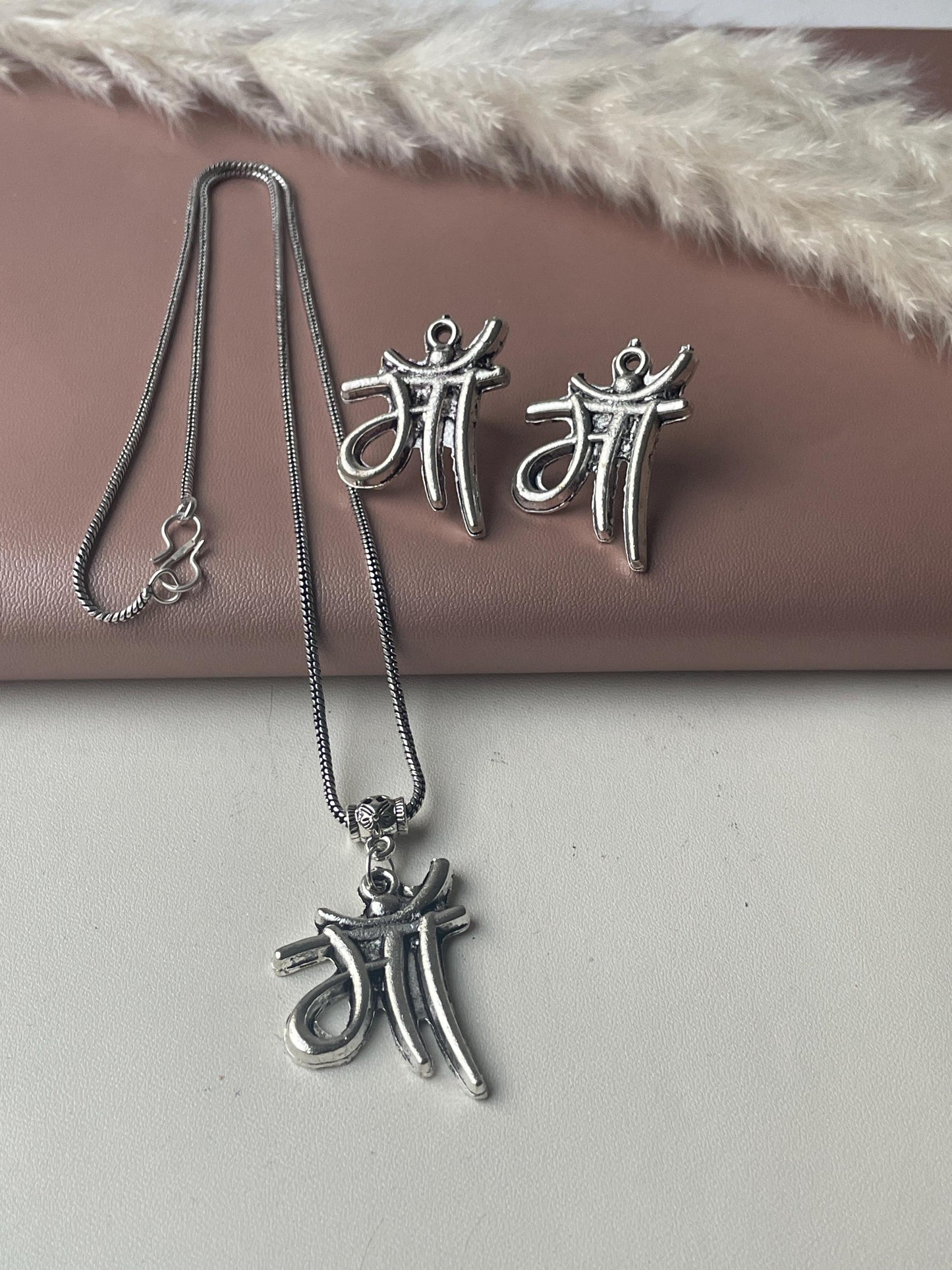 Silver necklace and earrings set. The pendant and earrings feature the Hindi word for mother, 'Maa,' in a stylized script. The necklace is a delicate chain with a small clasp. The earrings are small and lightweight. The jewelry has an antique silver finish. The set is displayed on a pink surface with a dried pampas grass decoration.