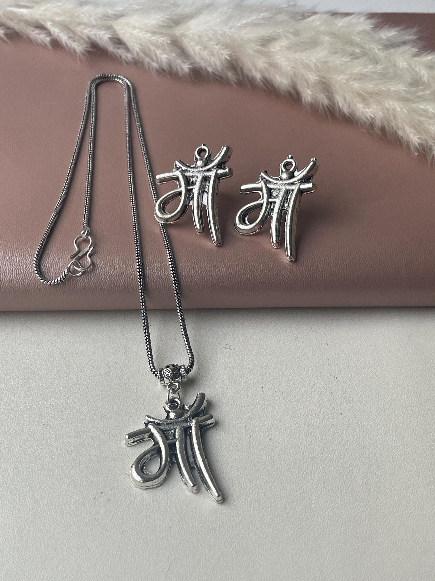 Silver necklace and earrings set. The pendant and earrings feature the Hindi word for mother, 'Maa,' in a stylized script. The necklace is a delicate chain with a small clasp. The earrings are small and lightweight. The jewelry has an antique silver finish. The set is displayed on a pink surface with a dried pampas grass decoration.