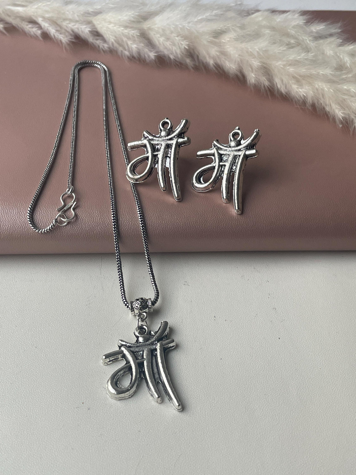 Silver necklace and earrings set. The pendant and earrings feature the Hindi word for mother, 'Maa,' in a stylized script. The necklace is a delicate chain with a small clasp. The earrings are small and lightweight. The jewelry has an antique silver finish. The set is displayed on a pink surface with a dried pampas grass decoration.