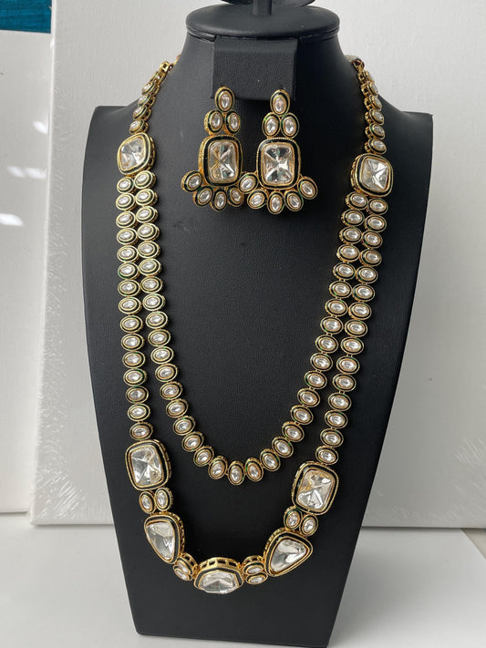 Gold plated kundan necklace and earrings set. The necklace is a double strand featuring clear cubic zirconia stones, kundan and dark green accents. The earrings are matching drop style. The jewelry is displayed on a black jewelry stand.