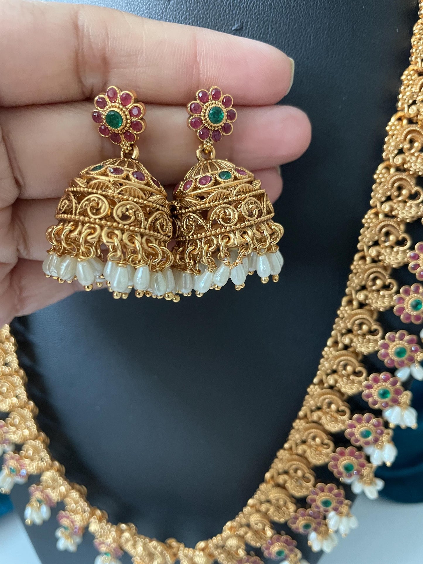 Gold necklace and earrings set. The necklace features a long chain adorned wpink and green floral accents and pearl drops. The earrings are jhumkas with similar pink and green floral detailing and pearl accents. The jewelry has an ornate, traditional design with intricate detailing.
