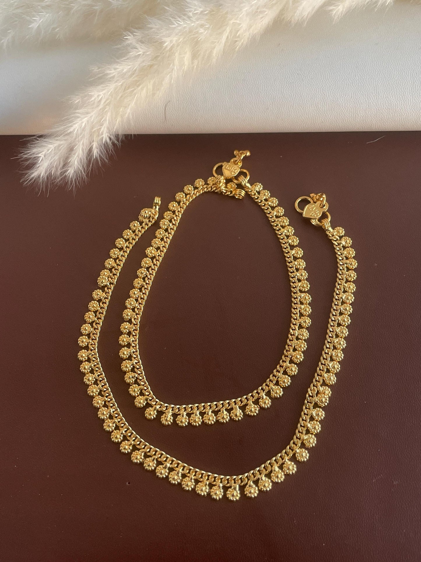 A pair of gold anklets with a delicate floral design. The anklets are made of a thin chain with small, round, flower-shaped charms.