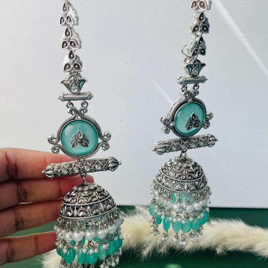 Silver-toned earrings with a long, ornate design. Each earring features a decorative top with a teal-colored, circular stone and a dangling bell-shaped bottom. The bell is adorned with intricate patterns and teal beads, along with white pearl accents.