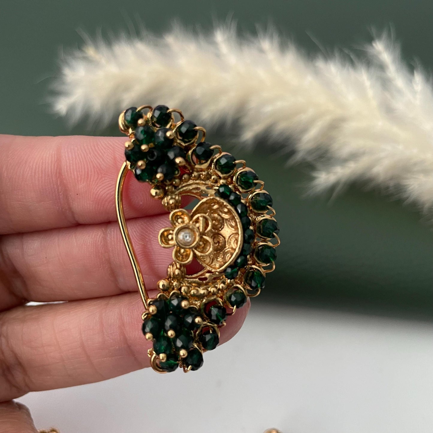 A gold-toned nose ring with a floral design and a crescent shape, adorned with dark green beads. The jewelry piece has a small, central flower with a single, clear gem.