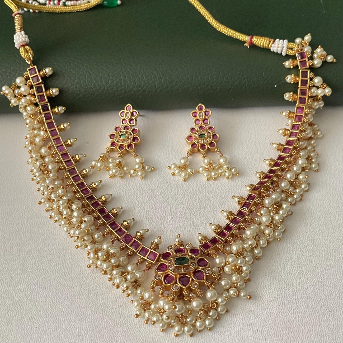 A gold-toned necklace and earring set. The necklace features a central floral design with a green stone, surrounded by red stones. The necklace and earrings have dangling white pearls. The necklace has a matching earring set.