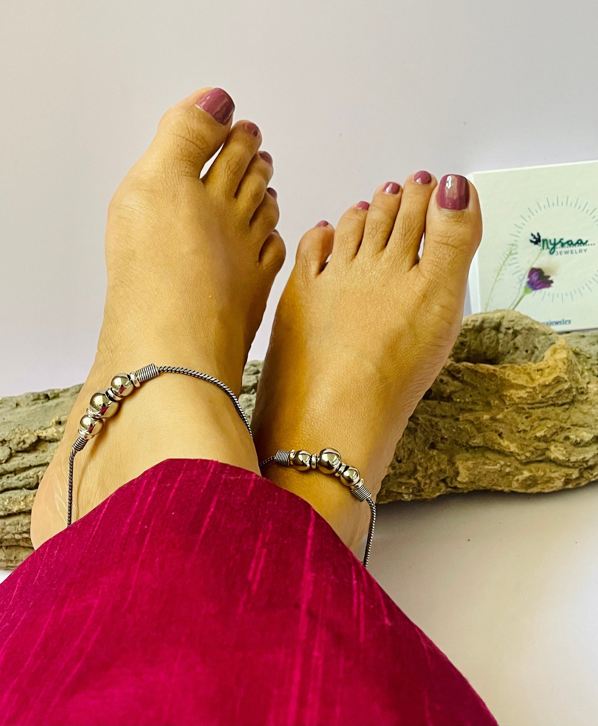Two silver anklets with small round beads. The anklets are on a pair of feet with pink nail polish.