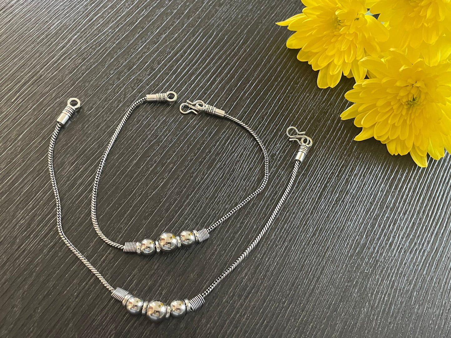 silver anklets with a chain design and a row of silver beads. The anklets are on a dark gray surface.