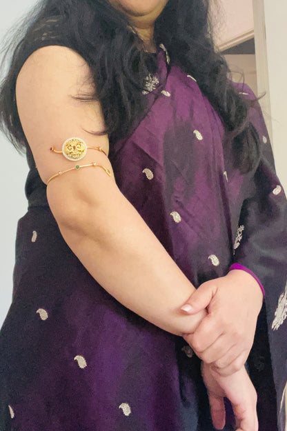 gold plated armlet with a round, ornate design and green stones. The armlet is worn on the upper arm.