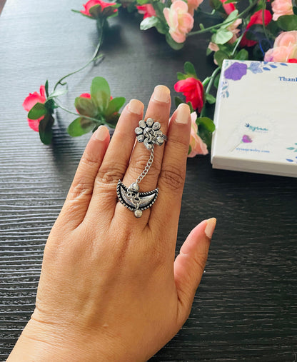 A silver ring with a flower design and a dangling crescent moon charm. The ring is worn on a finger and has a chain connecting the flower and moon.