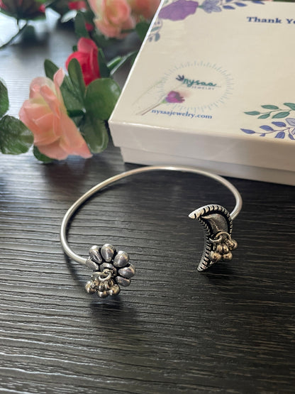 Silver bangle bracelet with a flower and crescent moon design. The flower has a small bell charm hanging from it. The crescent moon has a row of small bells hanging from it.