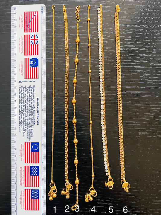 Six gold anklets with different designs. The anklets are laid out on a dark wood surface with a ruler in the background. The ruler is marked in inches and centimeters. The anklets are numbered 1 through 6.