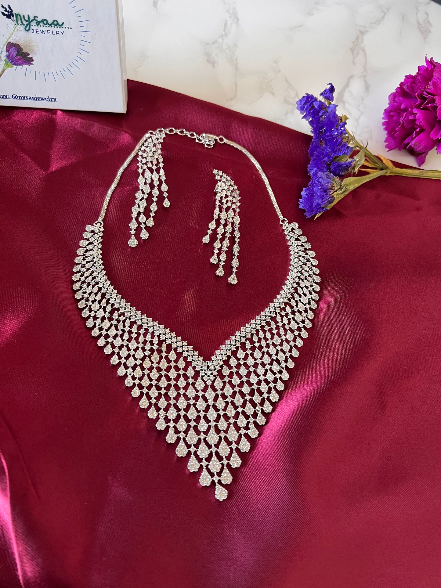 A rhodium plated CZ necklace and earrings set with a teardrop design. The necklace is a v-shaped design with multiple rows of teardrop-shaped cubic zirconia crystals. The earrings are long and dangle with multiple rows of teardrop-shaped crystals.