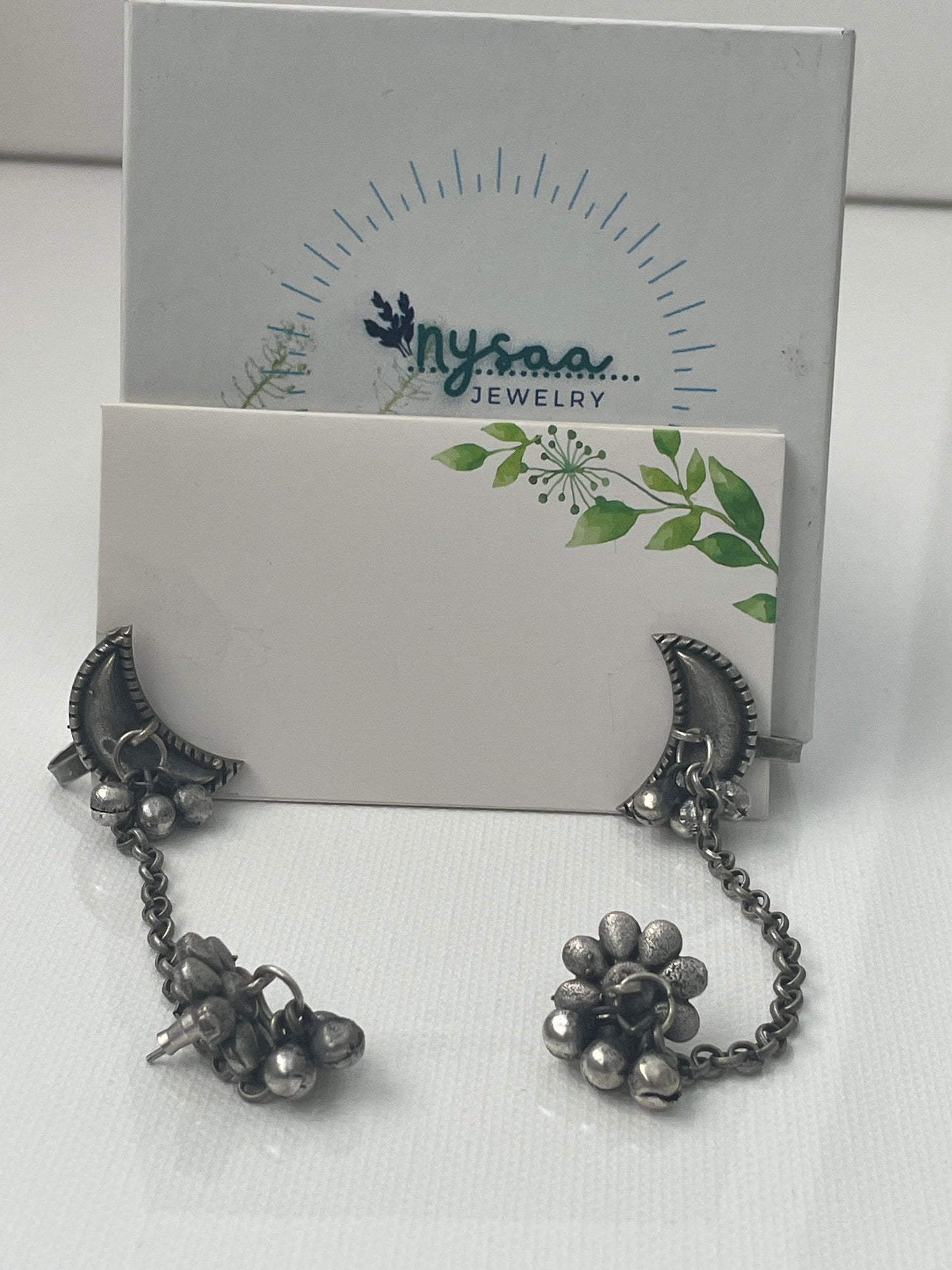 A pair of silver earrings with a crescent moon design and a dangling flower charm. The earrings are connected by a chain.