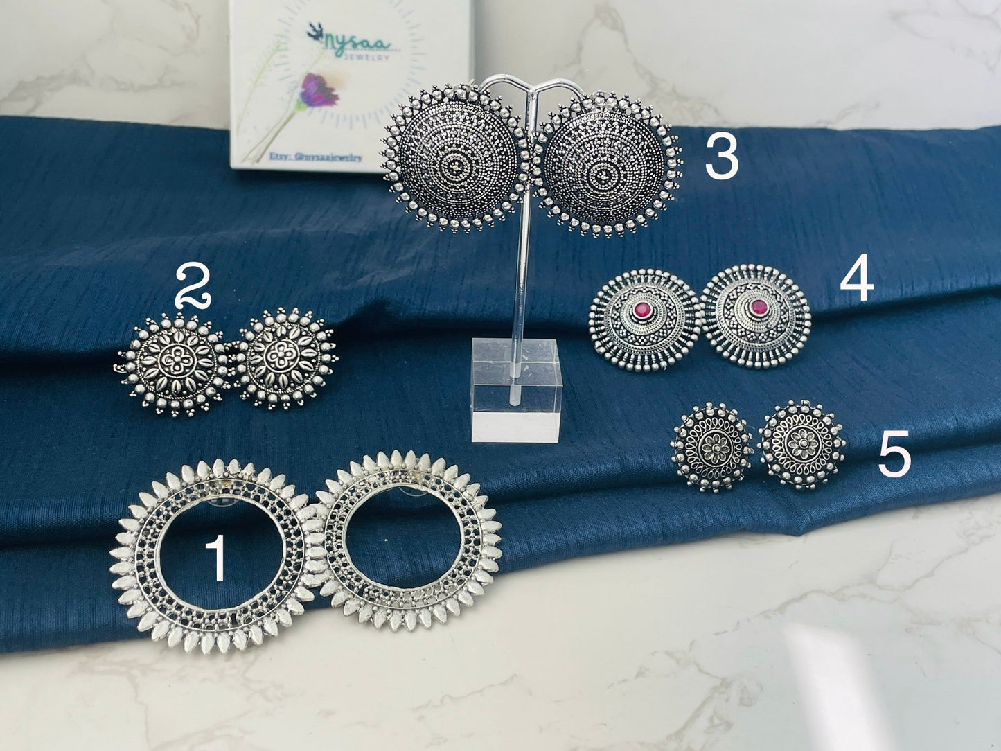Different designs of stud Oxidised earrings. The earrings are numbered 1 through 5. Earrings 1 are large and round with a circle in the center. Earrings 2 are round with a floral design. Earrings 3 are round with a mandala design. Earrings 4 are round with a floral design and a pink stone in the center. Earrings 5 are round with a floral design.