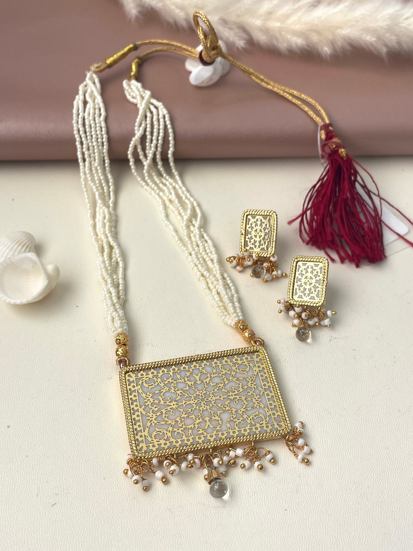 A gold and white thewa necklace and earrings set. The necklace has a rectangular pendant with a white center and intricate gold filigree design. The pendant is attached to a string of white beads. The earrings are also rectangular with a white center and gold filigree design. The earrings are attached to a string of white beads.