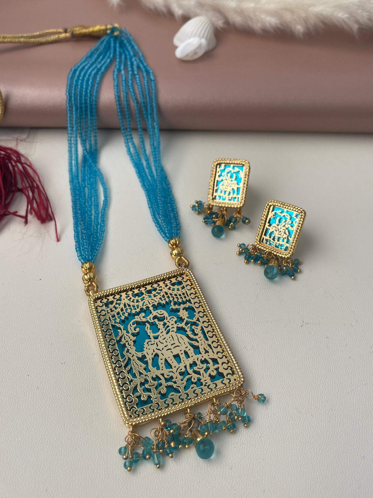 A gold necklace with a blue gold plated pendant featuring an intricate design of an elephant and people. The pendant is attached to a chain of blue beads. The necklace comes with matching earrings with a similar design.
