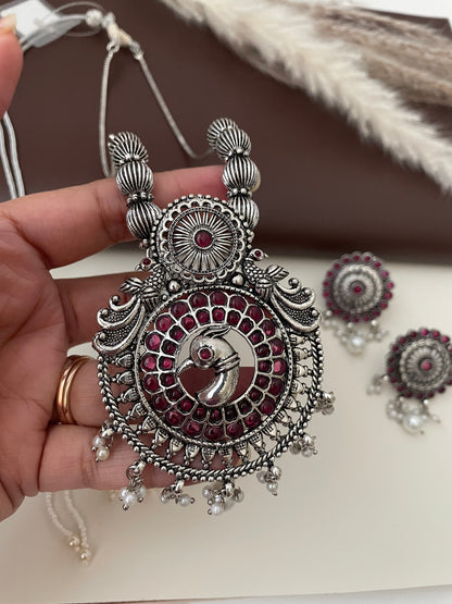 A silver necklace with a pendant featuring a peacock design with ruby kemp stones. The pendant is surrounded by a circle of smaller pink stones. The necklace has a silver chain and a silver tassel with small white pearls. The necklace is paired with matching earrings with a similar design.