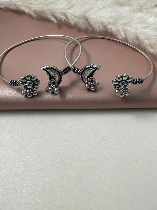 Two silver anklets with a crescent moon design and a floral design. The anklets are made of metal and have a silver finish.