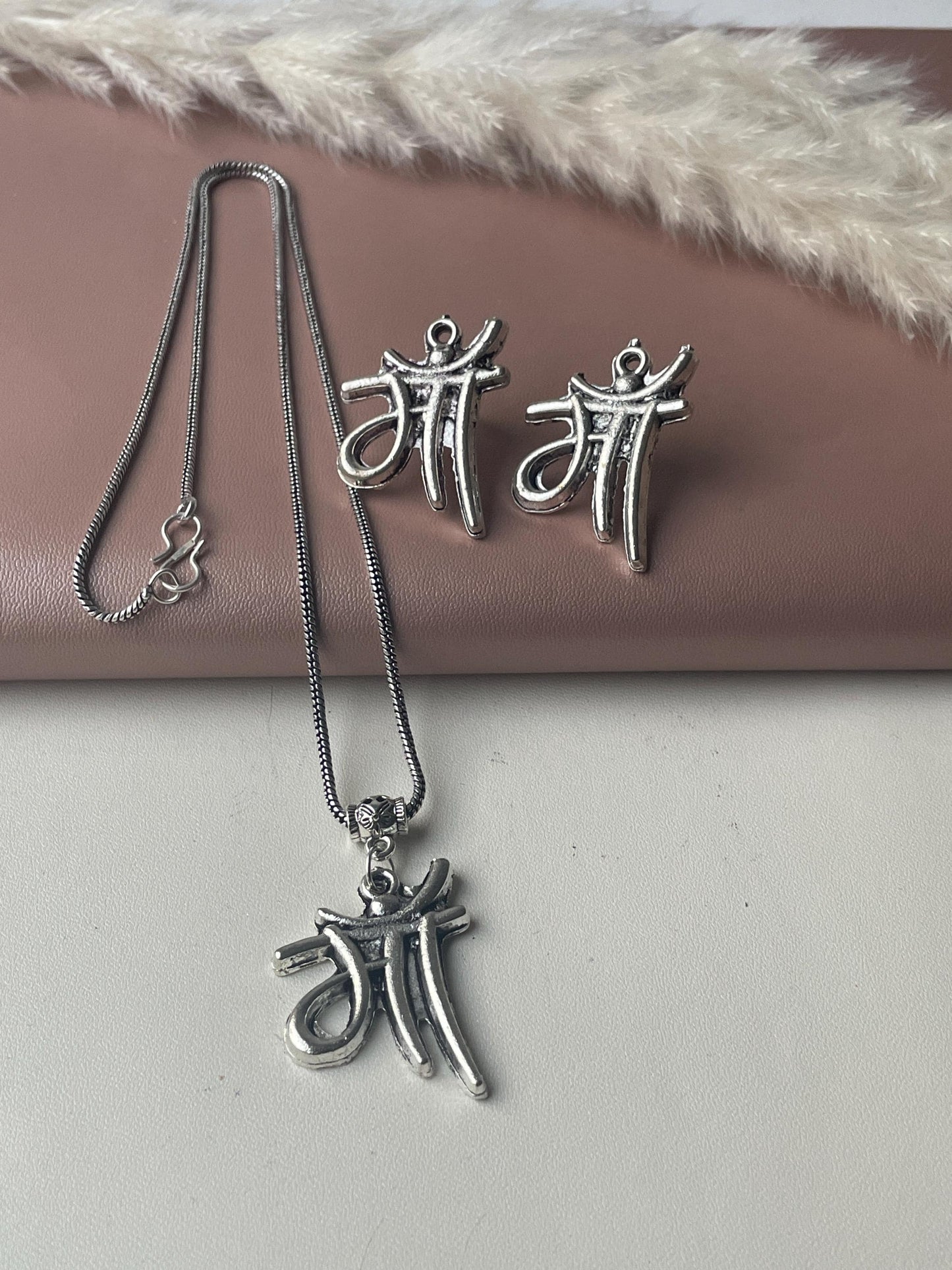 Silver necklace and earrings set.  The pendant and earrings feature the Hindi word for mother, 'Maa,' in a stylized script. The necklace is a delicate chain with a small clasp. The earrings are small and lightweight. The jewelry has an antique silver finish. The set is displayed on a pink surface with a dried pampas grass decoration.
