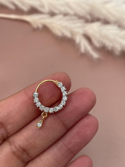 Gold nath nose ring with a circular design encrusted with small, clear cubic zirconia stones.  A single, dangling cubic zirconia stone hangs below the circular setting. The piece is delicate and sparkly, ideal for adding a touch of elegance to traditional attire.