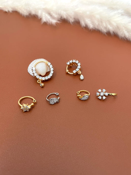 Six gold and silver nose rings with sparkling clear cubic zirconia nose pins.  Styles include a flower design, a curved shape, and a hoop with a dangling pear-shaped stone.  These nose rings are displayed on a brown surface.  The nose rings are small and delicate, perfect for adding a touch of sparkle to any look.