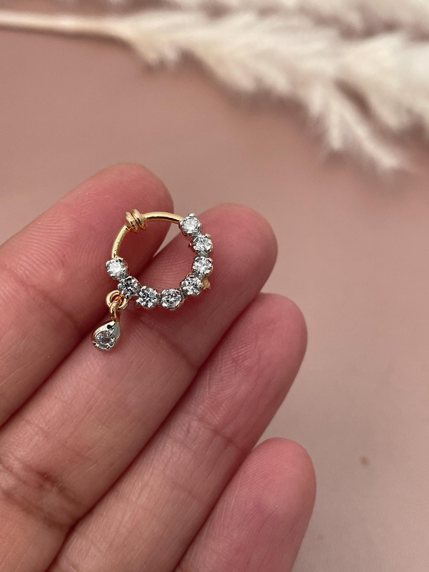 A gold nose ring with small, clear cubic zirconia stones. The nose ring is circular with a small dangling stone. The stones are prong-set and create a sparkly effect. The nose ring is delicate and elegant, suitable for everyday wear or special occasions.  It is a beautiful piece of jewelry that would make a great gift.