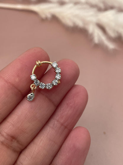 A gold nose ring with small, clear cubic zirconia stones. The nose ring is circular with a small dangling stone. The stones are prong-set and create a sparkly effect. The nose ring is delicate and elegant, suitable for everyday wear or special occasions.  It is a beautiful piece of jewelry that would make a great gift.