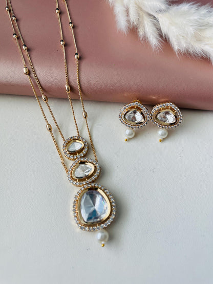 Gold necklace and earrings set. The necklace features three graduated pear-shaped clear stones framed by sparkling crystals, and a dangling pearl.  The matching earrings have a similar design with a single pear-shaped stone and pearl. The jewelry is displayed against a rose gold background.
