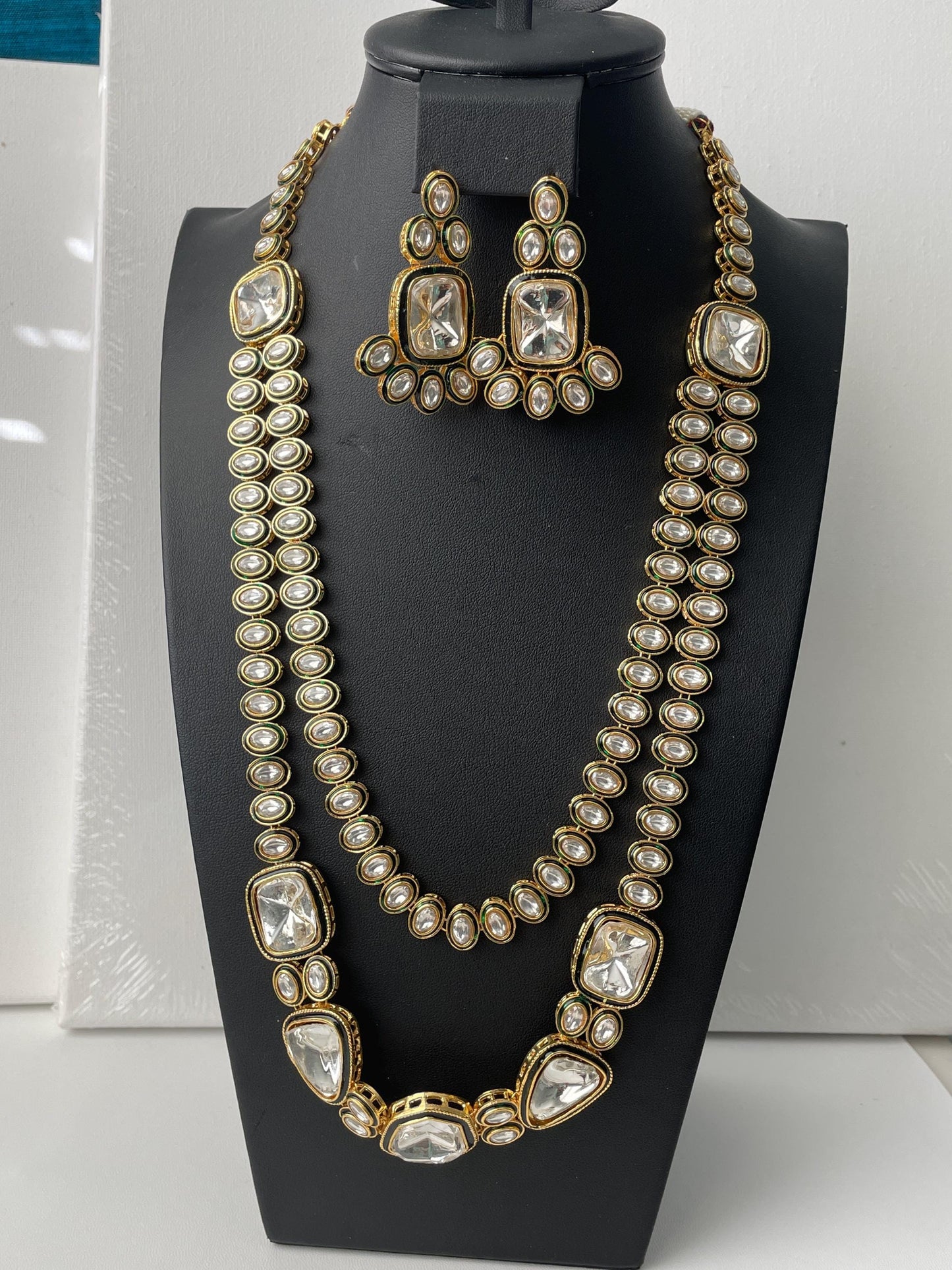 Gold plated kundan necklace and earrings set. The necklace is a double strand featuring clear cubic zirconia stones, kundan  and dark green accents. The earrings are matching drop style. The jewelry is displayed on a black jewelry stand.