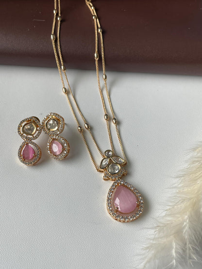 Gold necklace and earrings set. The necklace features a double chain with small beads and a pink pear-shaped pendant adorned with clear and pink stones. Matching earrings with pink stones and clear accents. The jewelry is elegant and delicate, perfect for adding a touch of sparkle to any outfit.