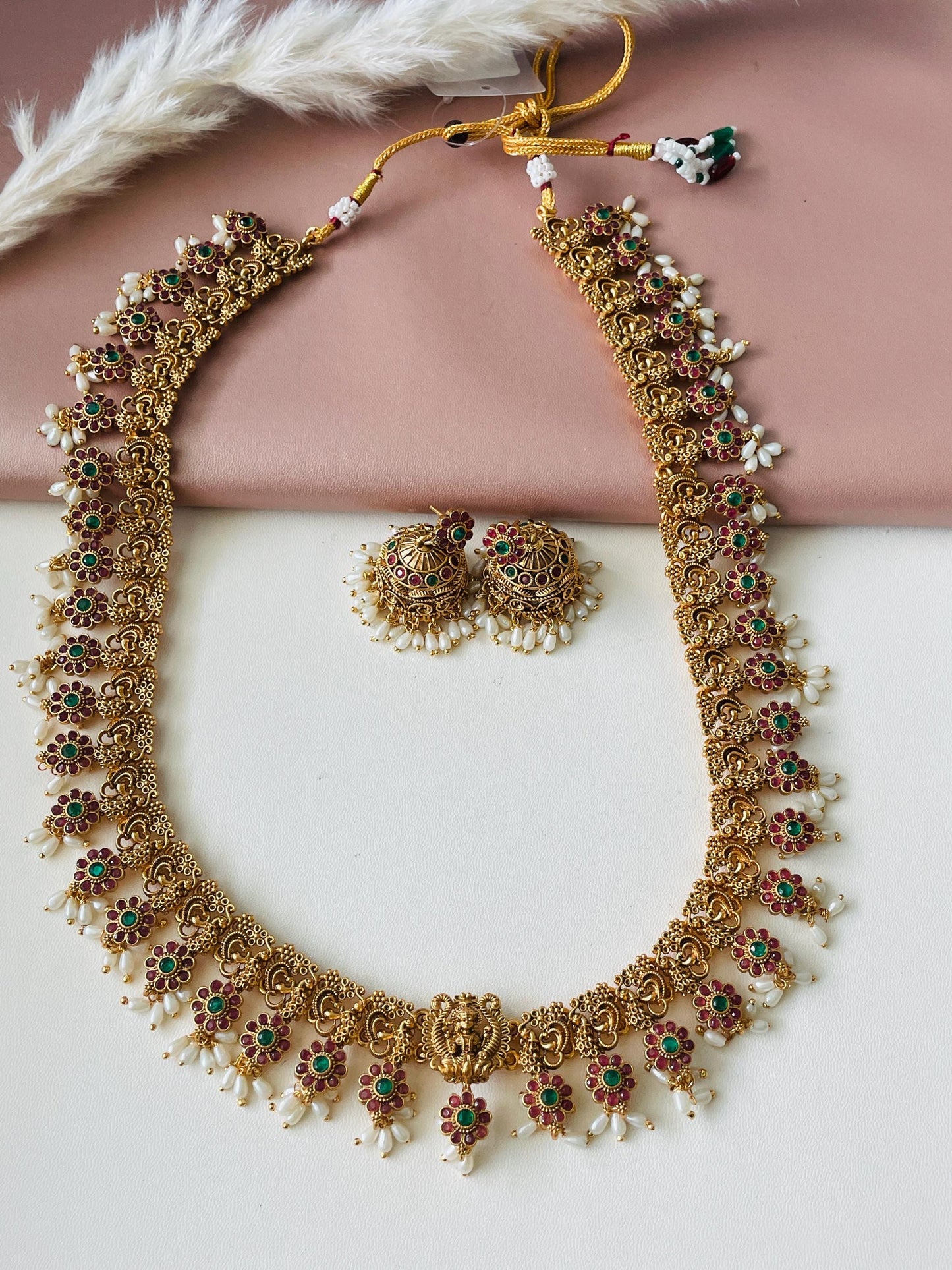 Gold necklace and earrings set. The necklace features a long, ornate chain with red and green stones, and pearl accents.  The matching earrings are jhumkas with similar red and green stones and pearls. The necklace has a traditional South Indian design. The jewelry is displayed against a pink background.