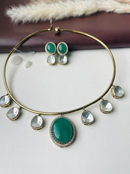 A gold choker necklace with a large green oval stone pendant surrounded by smaller clear stones. The necklace has a simple design with five smaller clear stones hanging from the choker. The necklace comes with a pair of matching earrings with green oval stones and smaller clear stones.
