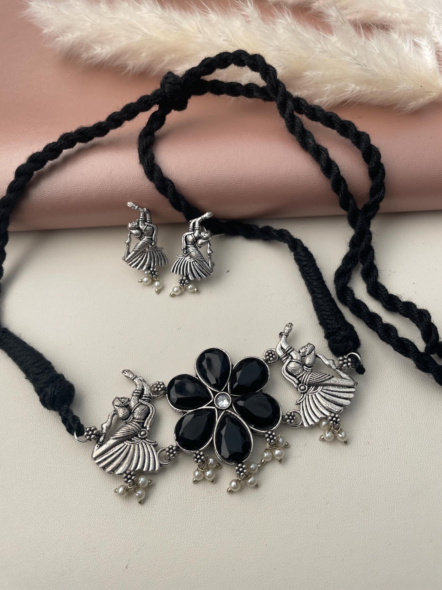 A silver necklace with a black flower pendant and two matching earrings. The pendant features a black stone surrounded by silver accents and small pearl-like beads. The earrings are also silver with a black stone and pearl-like beads.