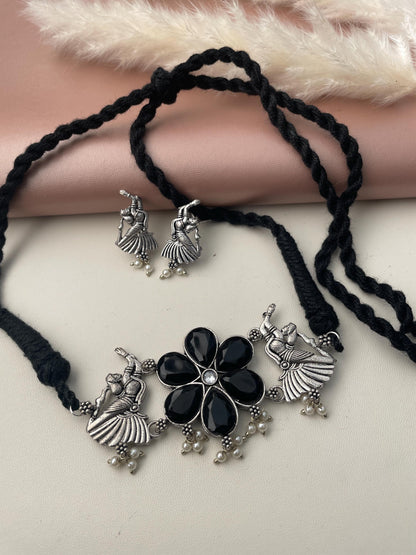 A silver necklace with a black flower pendant and two matching earrings. The pendant features a black stone surrounded by silver accents and small pearl-like beads. The earrings are also silver with a black stone and pearl-like beads.