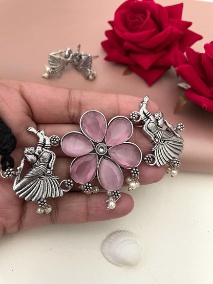 A silver necklace with a pink flower pendant and two silver figures of dancers. The necklace has a black cord and pearl accents.