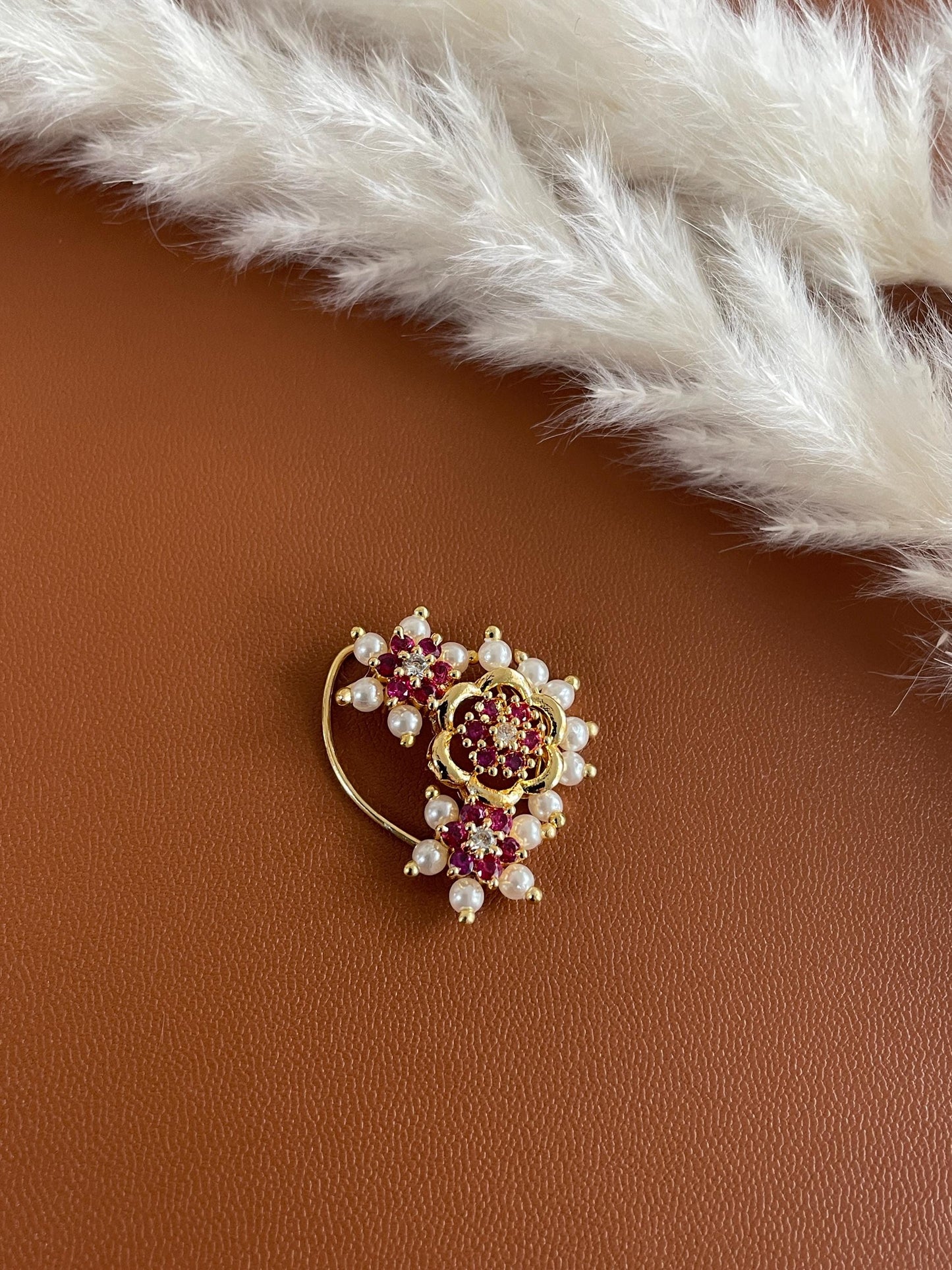 A gold nose ring with a floral design. The ring is adorned with red and white gemstones and pearls.