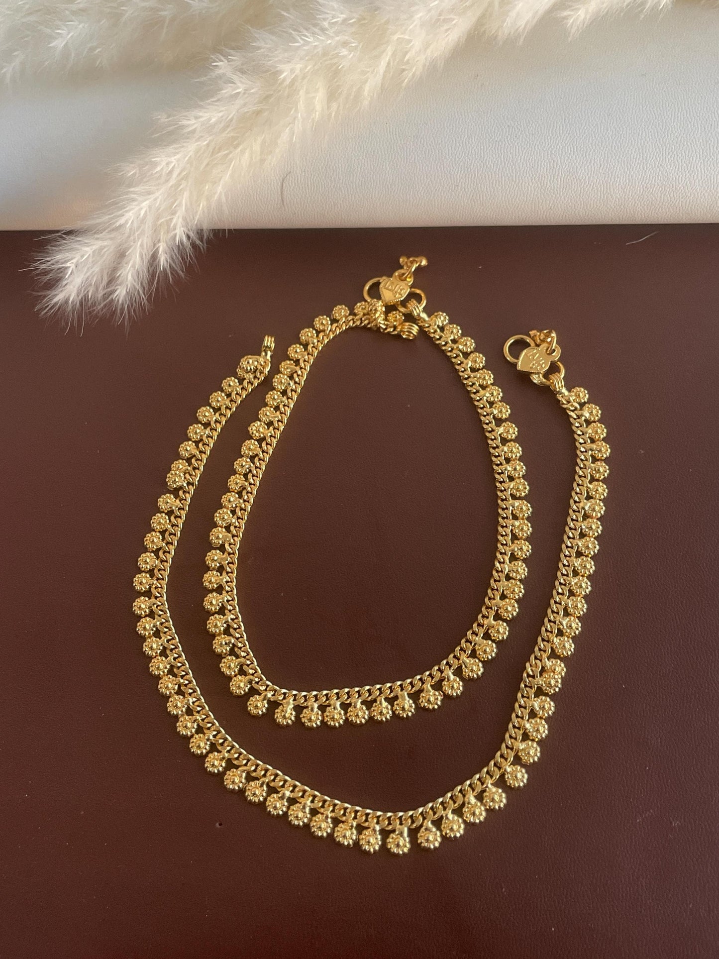 A pair of gold anklets with a delicate floral design. The anklets are made of a thin chain with small, round, flower-shaped charms.