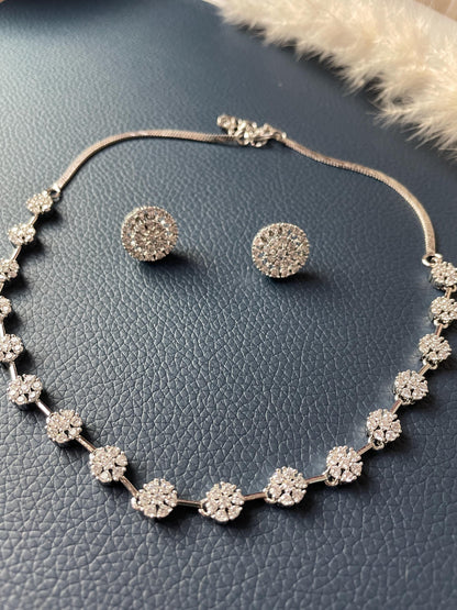 A silver necklace and earrings set with round, sparkly, white stones. The necklace has a chain with small, round, sparkly, white stones attached to it. The earrings are round, with small, sparkly, white stones ( cubic zirconia) attached to them. The necklace is rhodium plated and high quality.