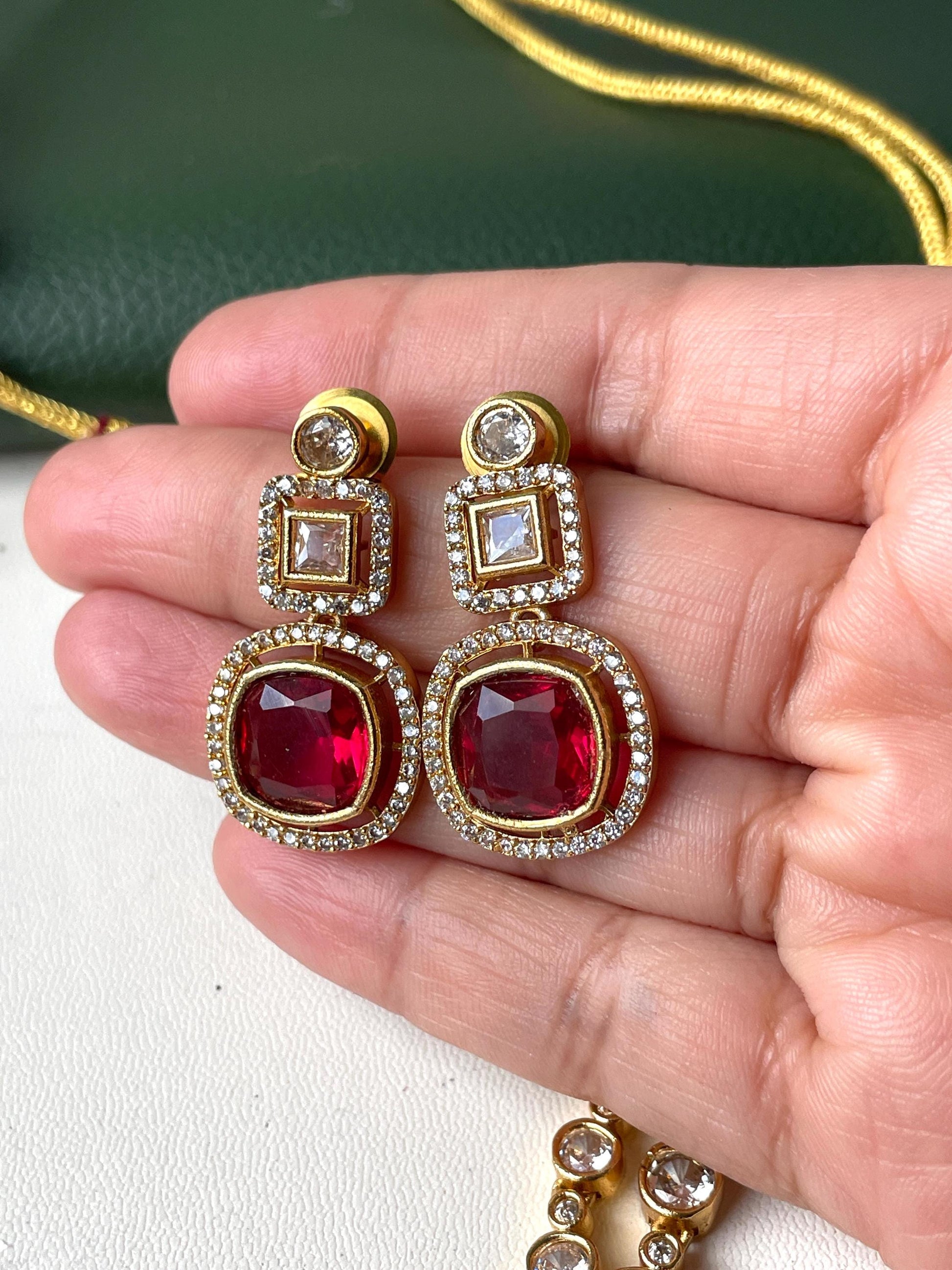 A pair of gold earrings with red and white stones. The earrings have a square shape with a smaller square in the center. The earrings are surrounded by smaller white stones.