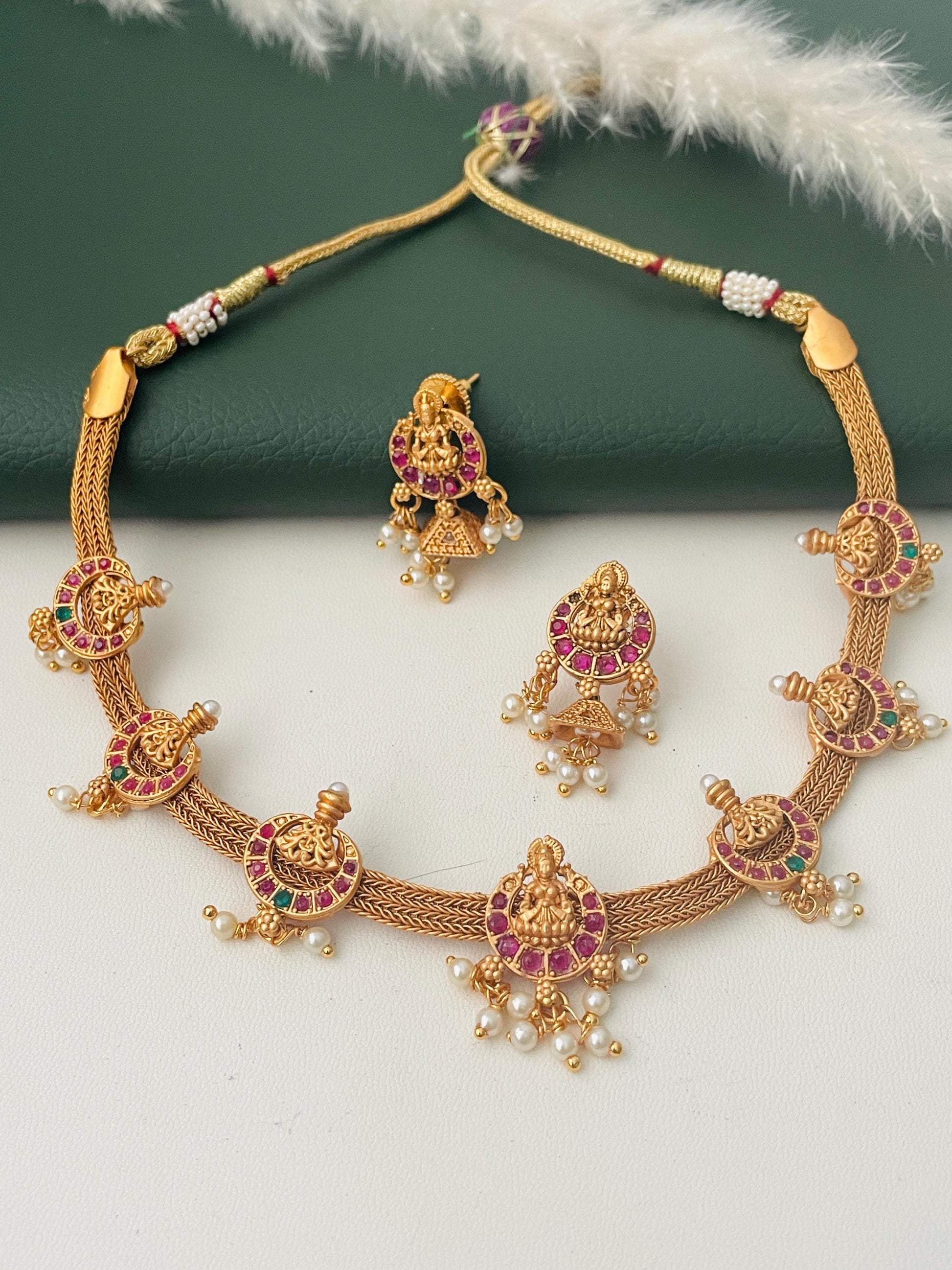 Stunning kemp stone gold plated short necklace set with matching earrings . This simple elegant yet very stunning necklace comes with small jhumkis. Lakshmi and flower motif withRuby and green kemp stones . 16 inches long necklace with adjustable dori .The South Indian temple themed jewelry.