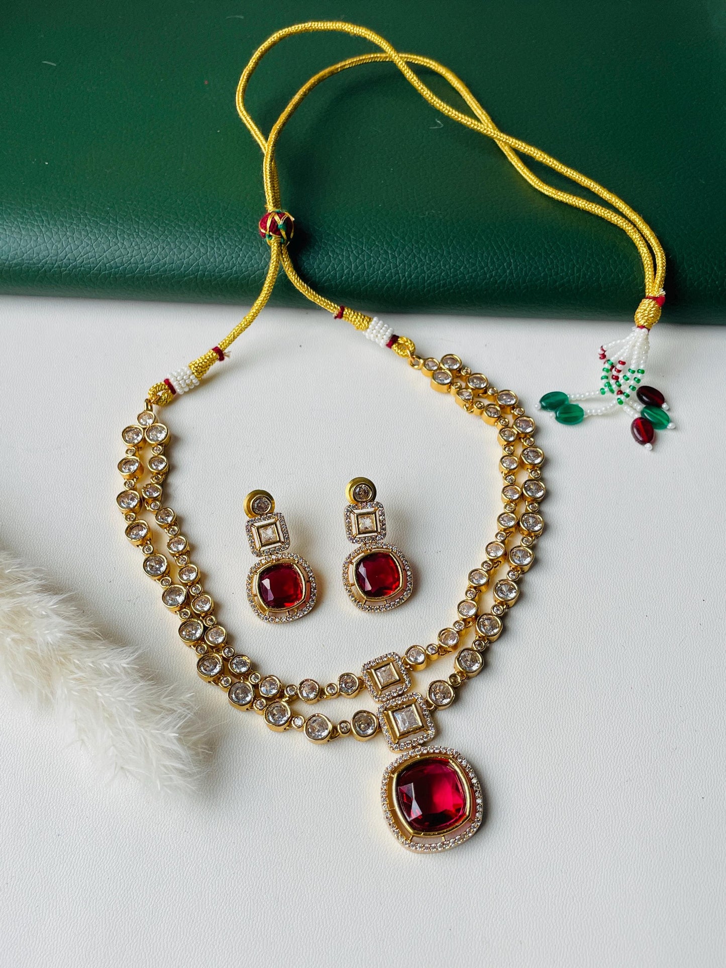 A gold necklace with a red gemstone pendant and matching earrings. The necklace has two strands of small clear stones and the pendant is surrounded by smaller clear stones. The earrings have a single red gemstone with clear stones around it.