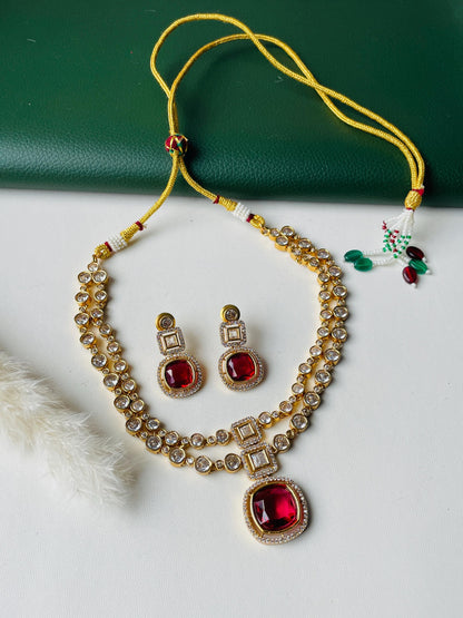 A gold necklace with a red gemstone pendant and matching earrings. The necklace has two strands of small clear stones and the pendant is surrounded by smaller clear stones. The earrings have a single red gemstone with clear stones around it.