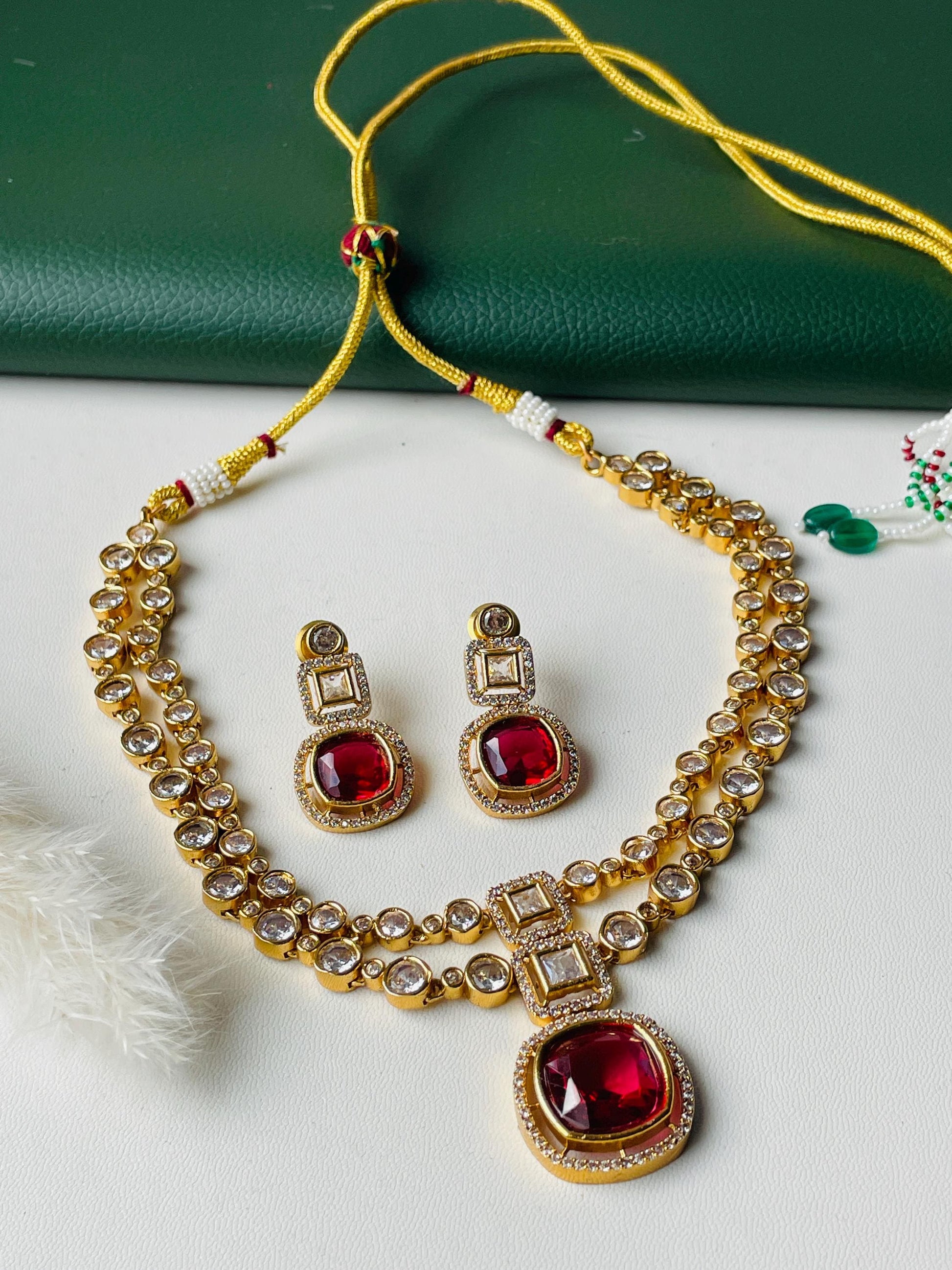 A gold necklace with a red gemstone pendant and matching earrings. The necklace has two strands of small, clear stones. The pendant is a square shape with a red gemstone in the center. The earrings are also square shaped with red gemstones in the center.