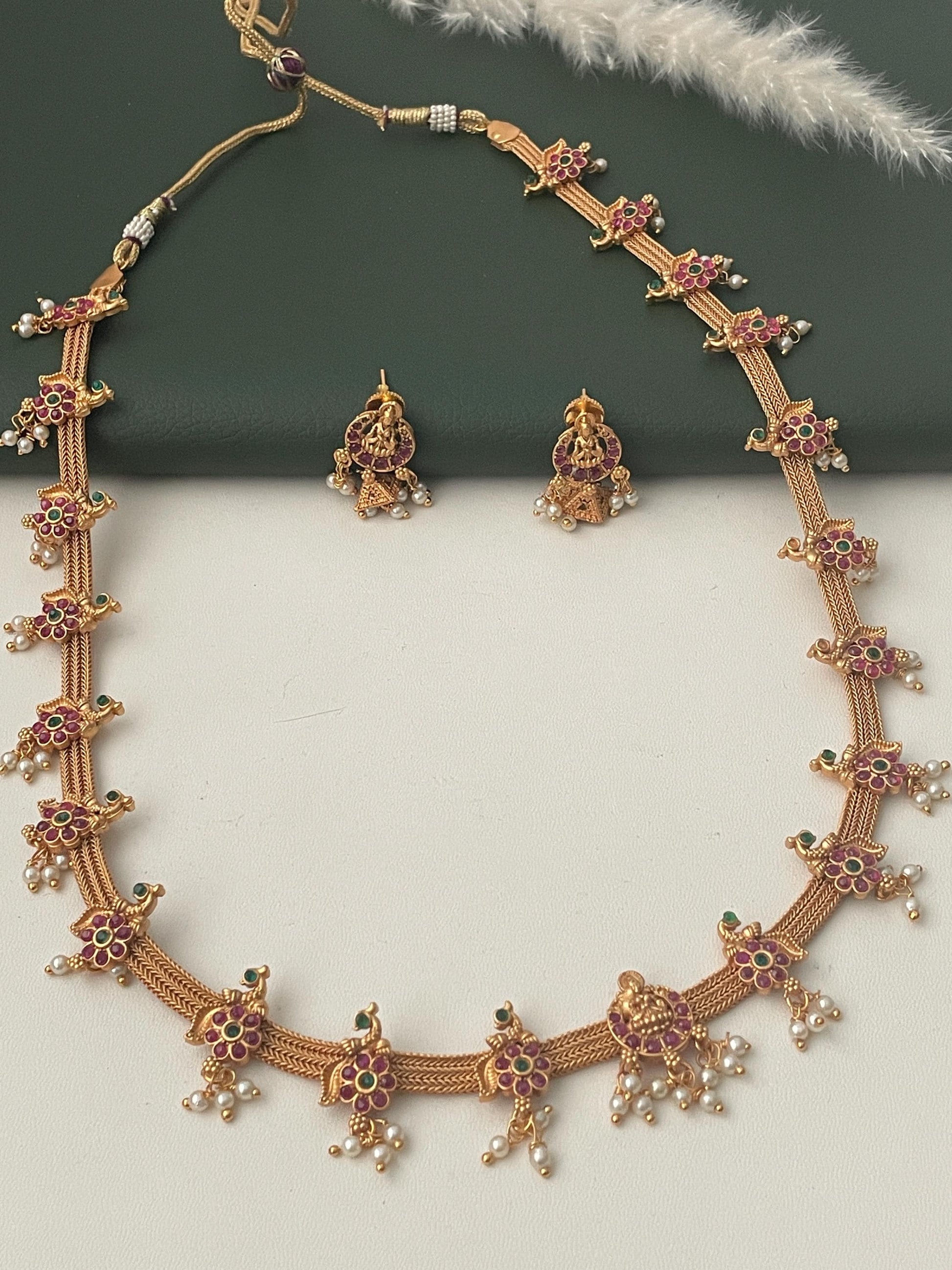 Stunning kemp stone gold plated long necklace set with matching earrings . This simple elegant yet very stunning necklace comes with small jhumkis. Lakshmi and flower motif withRuby and green kemp stones . 22 inches long necklace with adjustable dori .The South Indian temple themed jewelry.