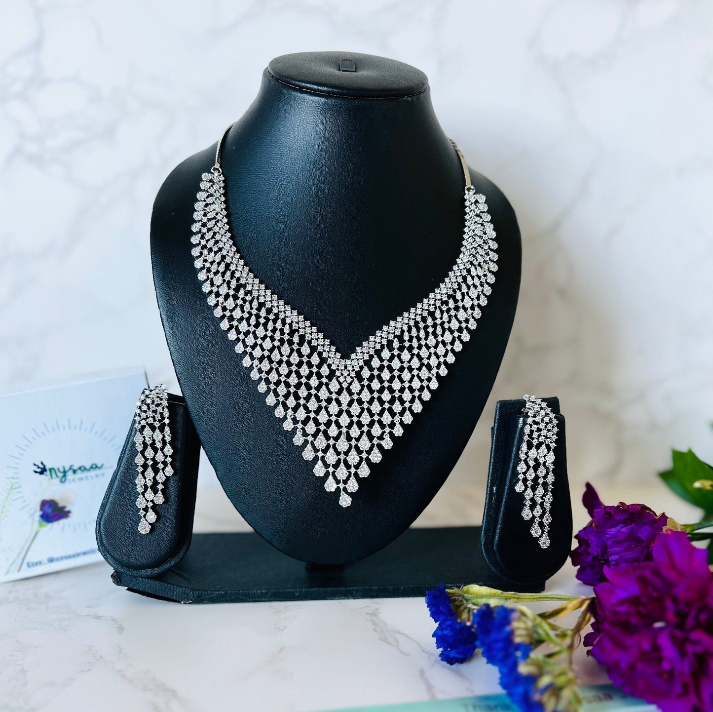 May include: A silver necklace and earrings set with a cascading design of small, clear stones. The necklace is displayed on a black stand and the earrings are displayed on a black stand with a white label that says 'nysaa jewelry'.