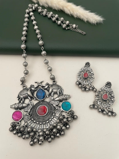 Oxidised necklace with a pendant featuring two elephants, a red, blue, and pink stone, and a silver chain. The necklace is paired with matching earrings with a red stone and silver chain.