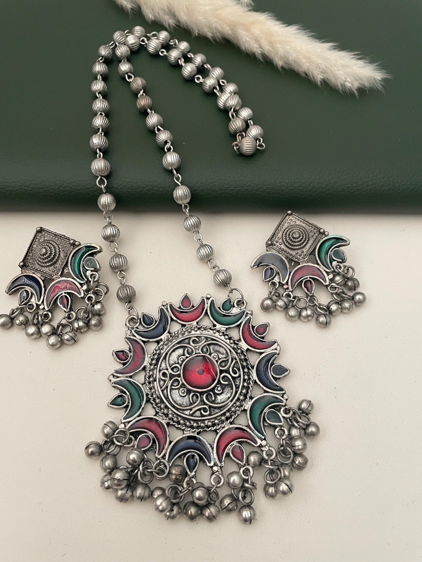 Oxidised necklace and earrings set with a large, round pendant. The pendant is decorated with a red, green, blue, and pink enamel design. The necklace and earrings have silver beads and dangling charms.